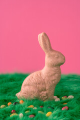 An Easter bunny made from swiss, white chocolate with raspberry chunks, sits on a green fur meadow with small colored Easter eggs made from sugar. Pink red background. Perfect for a easter theme