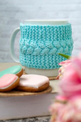 A white cup of tea in a knitted blue case stands on a book and pink flowers, tulips and gingerbread on a texture table. Spring concept