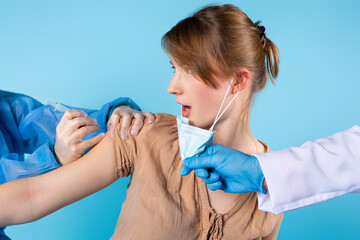 Cropped photo of a young blonde girl who looks to the side in surprise. The doctor's hands in latex gloves inject the vaccine into the forearm and remove the protective mask. Blue background.