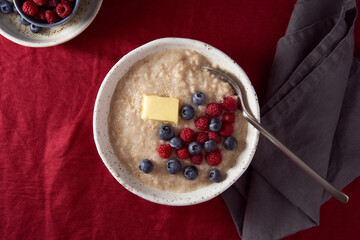 Oatmeal porridge with blueberry, bilberry. Breakfast in white bowl and spoon with fresh berries and butter on dark red cranberry linen tablecloth. Bright color of textile. Top view, copy space