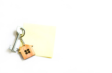 Keychain in the shape of wooden house with key on a white background with a square sheet for notes. Building, design, project, moving to new house, mortgage, rent and purchase real estate. Copy space
