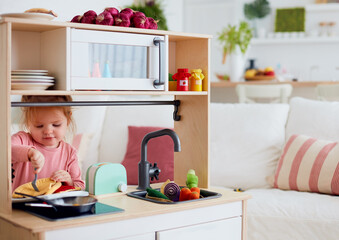 cute toddler baby girl playing on toy kitchen at home, pretends eating pizza with a fork