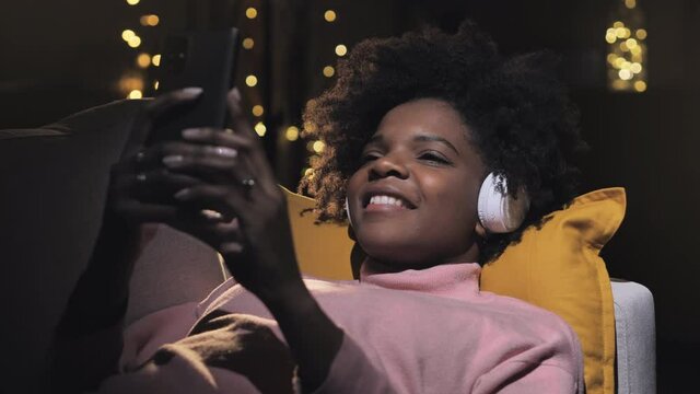 Carefree smiling black young woman listen to music lying on sofa at home using smart phone in a dark room,relaxed girl wearing headset holding telephone looking at the screen enjoy playing song