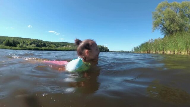 A happy little girl is floating on the river in inflatable armbands, on a bright sunny day, against a background of blue sky and green reeds. Close-up. View from the water surface. 4K