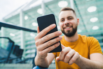 Young handsome smiling bearded man sitting at stadium steps and making bets online on his...