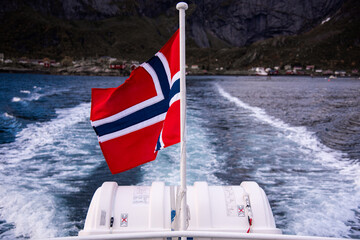 Norwegian Flag waving in the wind on a boat with landscape with Reine fjord, Lofoten Islands, mountains, forest and glacier in Norway - the photo was taken from a boat - sunshine and light rays
