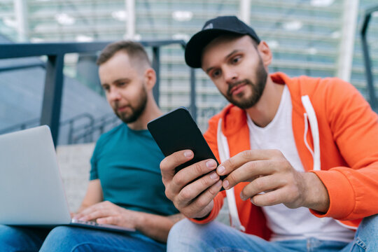 Two friends sitting on football stadium steps and making bets using smartphone and laptop