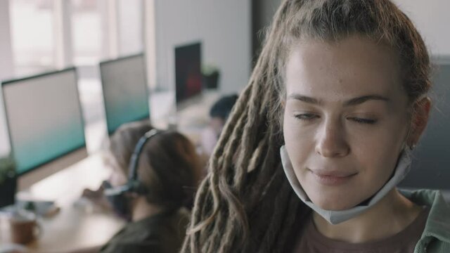 Portrait close up with PAN of young woman with dreadlocks wearing face mask under her chin and posing for camera. Her colleagues in headsets working on computers in background