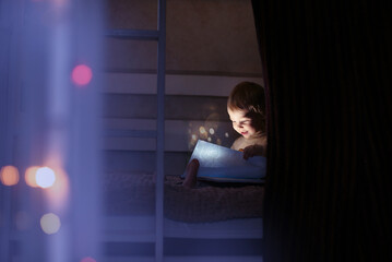 a little girl is sitting on the first floor of a two-story bed in the dark, holding a book of fairy tales, looking at it and smiling