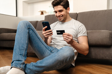 Happy guy using mobile phone and credit card while sitting on floor