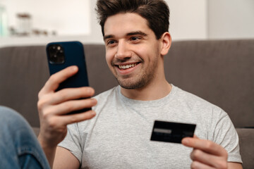 Happy guy using mobile phone and credit card while sitting on floor