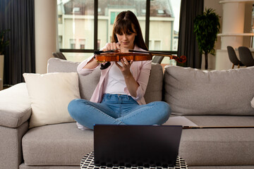 Young woman having online lesson of violin via laptop at home.