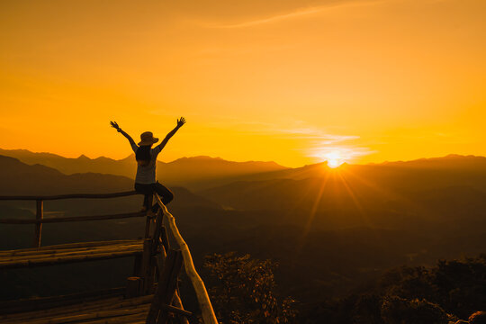 Picture from the back of a woman sitting and raise both arm on a wooden porch extending into a high mountain cliff. The sun is setting on the mountain and there is a beautiful warm orange light.