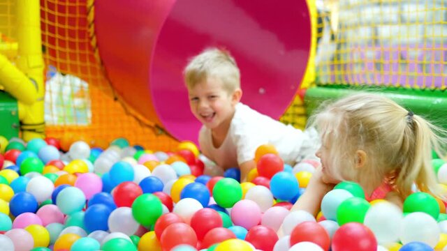 Having fun in playroom. Children boy and girl playing on indoor playground in multi colored plastic balls in big dry paddling pool in playing centre down slide and swing. Leisure Activity.