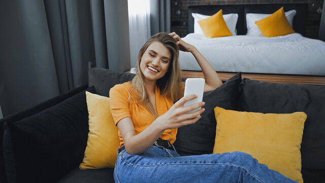cheerful woman taking selfie on couch in hotel room