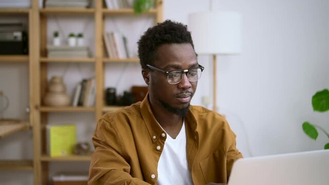 Businessman working with laptop and sitting at table at home during pandemic spbas. Young American man looks at computer screen and types, sits at desk in light interior. Millennial African