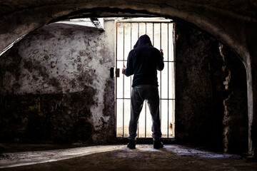 Prisoner man locked up standing in old underground cellar , silhouette from behind against bars -...