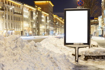 Vertical billboard on the sidewalk next to a snowdrift. The historic center of the evening city with illuminated facades. Mock-up.