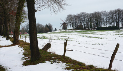 The old windmill on the hill is seen in the background. A snow covered meadow, barbwire fence and some trees in front.  A scenic winter landscape in Germany near the Dutch border.
