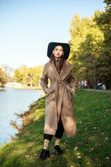 Stylish young lady in long beige coat and black hat walking by the river in city green park