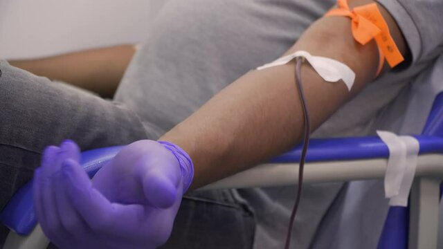 Side close-up, panning shot of a man arm while opening and closing her hand with blue gloves while donating blood with a catheter attached with medical tape.