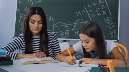 mother looking at book near daughter writing on notebook while doing homework