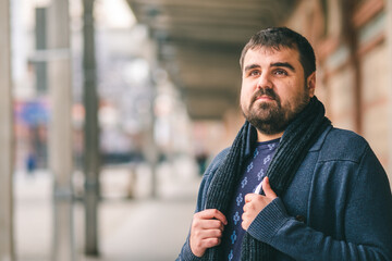 Bearded guy in blue sweater standing on the street with his hands on his scarf, looking up. 