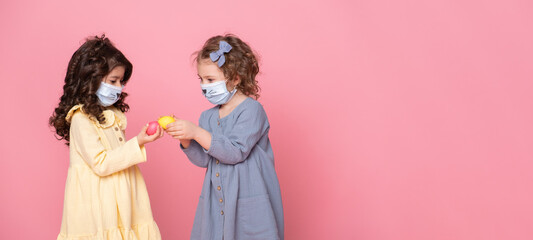 two girls in protective mask with colored eggs on pink background. Covid easter concept. Banner copyspace.