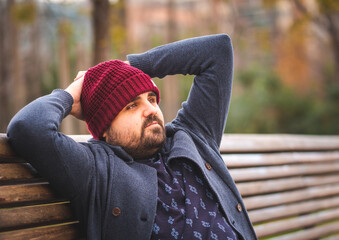 Boarded guy in a woolen hat and sweater sitting on park bench relaxing. 