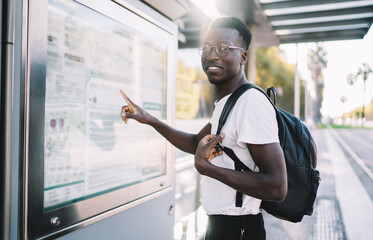 Handsome dark skinned male traveler with backpack checking location and destination on map, positive african american hipster guy checking public transport route on info banner on bus stop in city