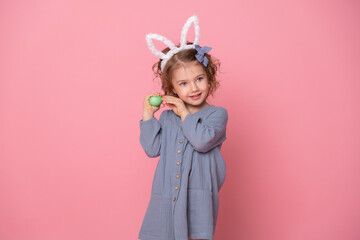 Obraz na płótnie Canvas Funny cute child girl in Easter bunny ears and blu dress holding colorful eggs on pink background