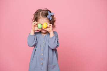 Funny cute child girl in blu dress holding up a colorful Easter egg in front of her eye on pink background