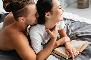 Shirtless man touching neck of cheerful girlfriend near book on blurred background