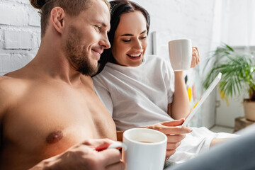 Sexy man holding digital tablet near cheerful woman with cup