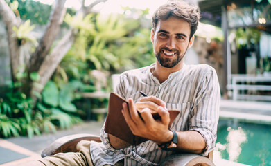 Fototapeta premium Happy man writing in notebook near swimming pool