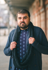 Bearded guy in blue sweater, standing on the street with his hands on his scarf, looking at the camera. 