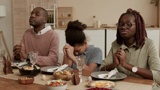 Medium close-up of African American family sitting at table in dining room with eyes closed and holding palms of hands in prayer before Easter dinner