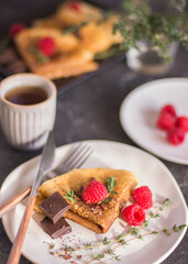 thin homemade pancakes with berries and chocolate on a dark background on a light plate, with herbs and raspberries and tea, close-up, vertical arrangement
