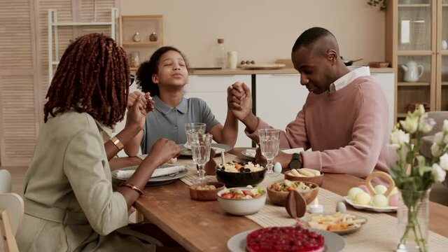 Happy Black family sitting at Easter dinner table, daughter taking hands of mom and dad, they closing eyes and starting praying