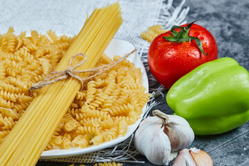 Raw spaghetti and fusilli on a white plate with tomato, pepper, and garlic