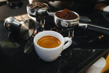 Espresso shot in white glass and tamper, filter with coffee beans, port filter on black table top. 