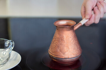 A copper turk for brewing coffee stands on a glass-ceramic plate. A woman's hand holds the handle of a turk. Make coffee in a turk.