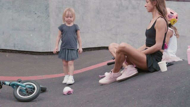 Wide shot of young woman and little girl sitting on asphalt in outdoor skating arena and watching little blond-haired girl approaching with bike and then throwing it on the ground