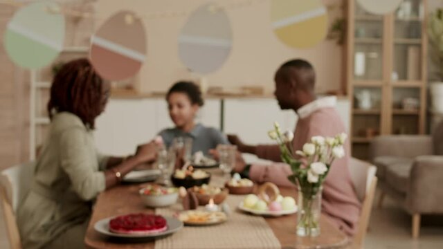 Medium long shot of happy Black family sitting at table, talking, having festive dinner, focus racking to decorative garland of paper Easter eggs hanging in room