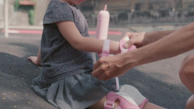 Close up of little Caucasian girl wearing skirt, t-shirt and white sneakers sitting on asphalt while her unrecognizable mommy helping her putting on wrist pads