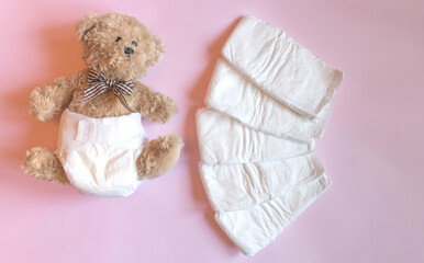 White disposable diapers for little babies on pink background and cute teddy bear. Top view,...