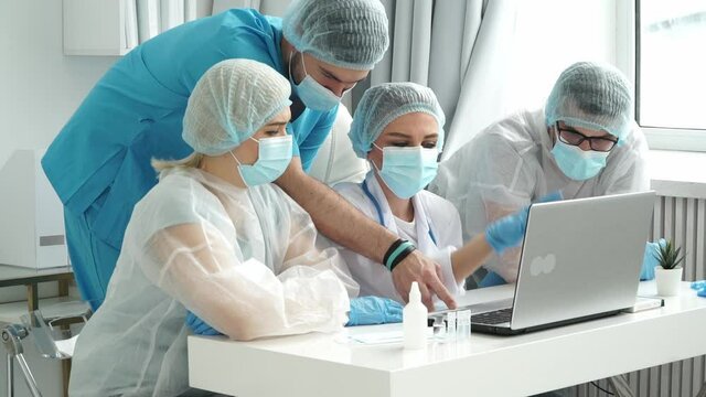 Group of doctors in medical gloves, white coats and protective masks analyze patients diagnosis at laptop in clinic. Treatment concept for coronavirus, covid-19