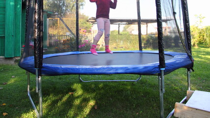 One little Russian girl jumps on a outdoor 8ft trampoline with outside safety soft net fence on green grass in the rural Garden at Sunny summer day, active sport vacation jumping