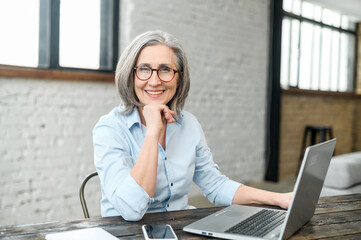 Intelligent elderly gray-haired businesswoman using a laptop in the office. Smart mature woman entrepreneur looks at the camera sitting at the desk. Senior online teacher, elderly female employee 