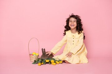 funny child girl in yellow dress with rabbit, tulips, basket with colorful eggs on pink background.
